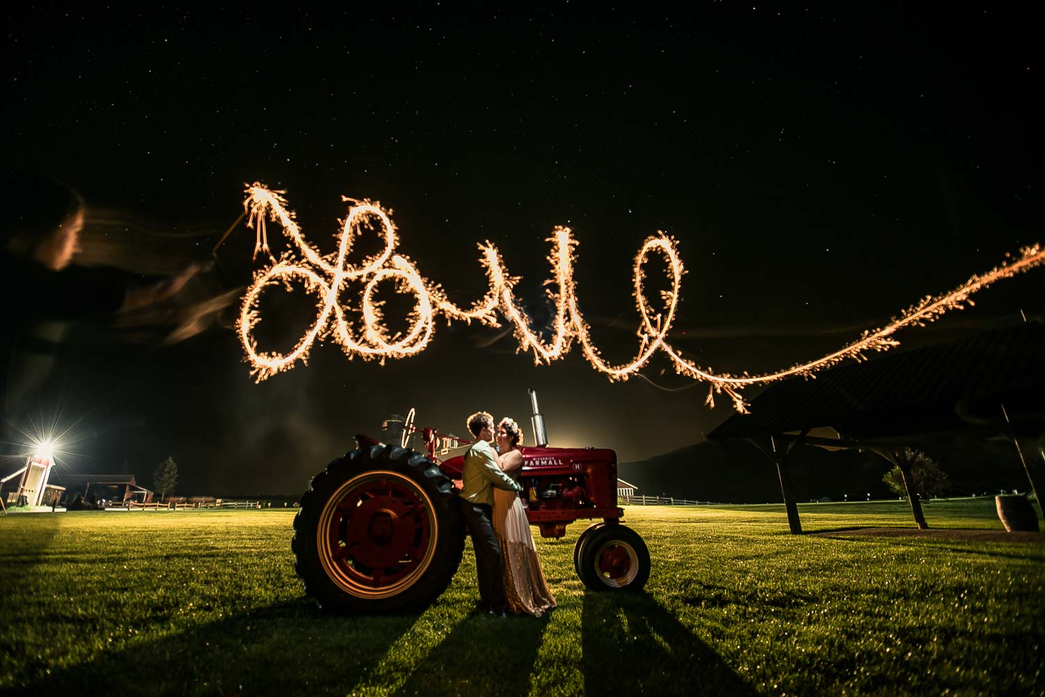 Wedding couple stands by tractor at night at Boyden Farms in Cambridge Vermont while sparkler writes the word love