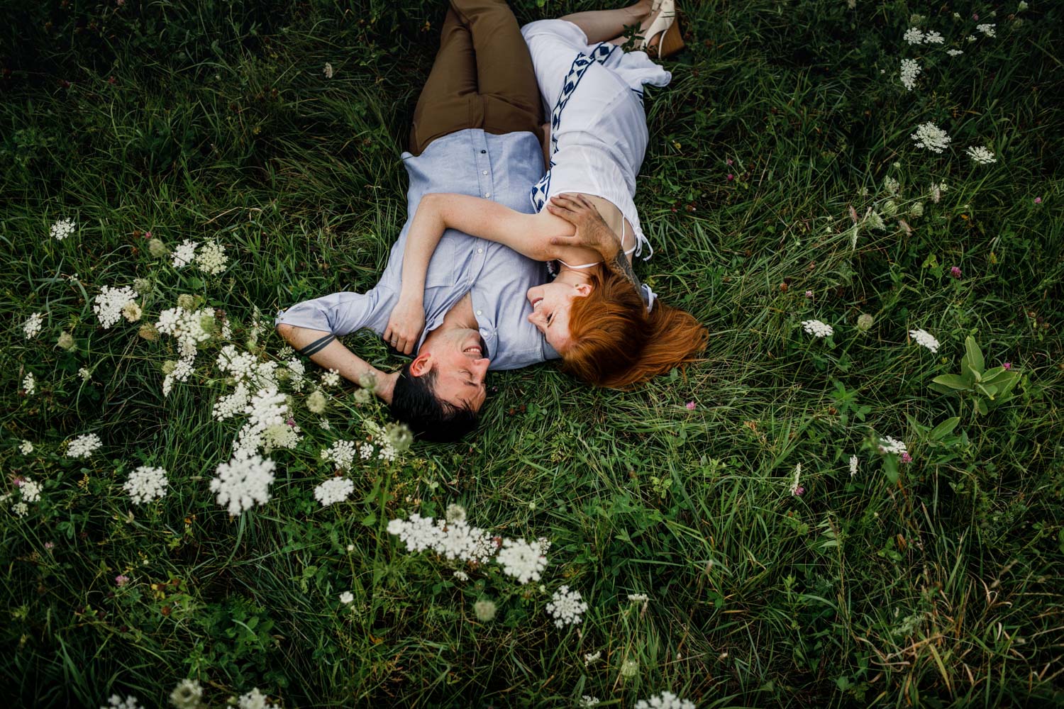 Engaged couple during Vermont wedding photographer engagement photo session lies in tall grass with flowers embracing in Huntington